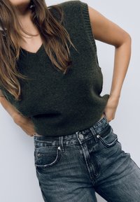 Temnozelena pletena brezrokavnik z V-izrezom in kratkimi rokavi, v kombinaciji z visokim pasom ter bledimi črnimi kavbojkami z srebrnimi detajli.