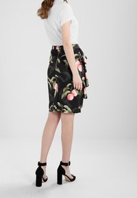 Žena v bílém tričku s krátkým rukávem, černé sukně ke kolenům s růžovým květinovým vzorem a černých sandálech na vysokém podpatku stojí bokem.