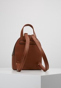 Mochila de cuero marrón con forma redondeada, doble asa superior y correas ajustables para los hombros. Presenta detalles en hardware dorado y un acabado texturizado.