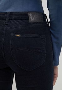 Jeans de pana oscuros con un bolsillo trasero bordado, una etiqueta de cinturón de cuero negro y un acento de logo dorado "Lee".