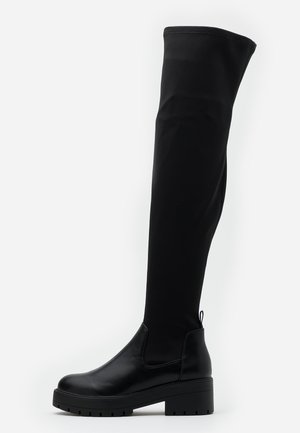 Botte noire au-dessus du genou avec semelle épaisse, talon bas et tige extensible sur fond blanc.