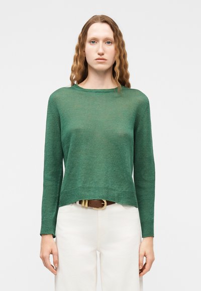 Femme aux cheveux roux ondulés portant un pull vert à manches longues, un pantalon blanc et une ceinture marron, se tenant devant un fond clair uni.