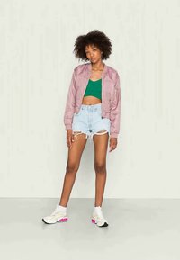 Jaqueta bomber rosa, top crop verde, calções de denim desgastados, ténis brancos com destaques em rosa. Modelo está de pé contra um fundo neutro.