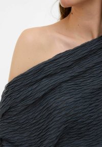 Mörkgrå, texturerad sjal med ett rynkat mönster som hänger över axeln. Tyget verkar mjukt med en lätt glans. Slät halsringning synlig.