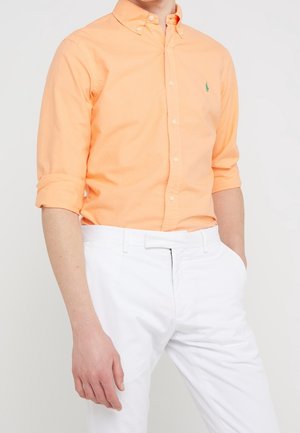 Mężczyzna w brzoskwiniowej koszuli z podwiniętymi rękawami i białych spodniach, z jedną ręką w kieszeni, na jednolitym tle.