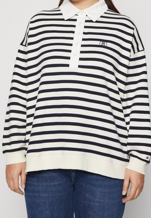 Person trägt ein weißes langärmeliges Polohemd mit horizontalen schwarzen Streifen und blauen Jeans, mit einem kleinen Logo auf der Brust und am Ärmel.