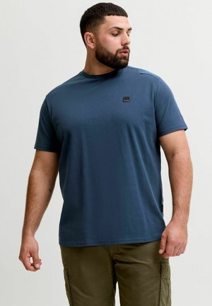 Uomo con la barba che indossa una camicia blu a maniche corte e pantaloni cargo oliva, guarda verso sinistra contro uno sfondo chiaro e uniforme.