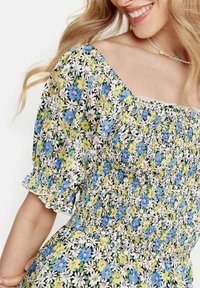 Meikkeis ommeltu kukkamekko, jossa on neliön muotoinen pääntie, puffimaiset hihat sekä monivärinen kuosi, jossa on sinisiä, keltaisia ja valkoisia kukkia mustalla taustalla.