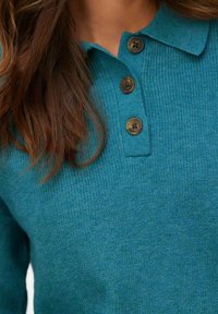 Laguna zielona dzianinowa koszulka polo z trzema brązowymi guzikami, z kołnierzykiem i prążkowaną fakturą. Zbliżenie ukazuje szczegóły tkaniny.