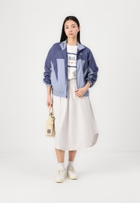 Windbreaker jacket in navy and light blue, with elastic cuffs; paired with a white shirt and beige skirt. Accessories include a small beige bag.