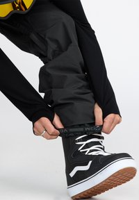 Botte de snowboard noire avec des accents et des lacets blancs, portée avec un pantalon noir ample. Le bas du pantalon a une bande élastique réglable pour un ajustement optimal.
