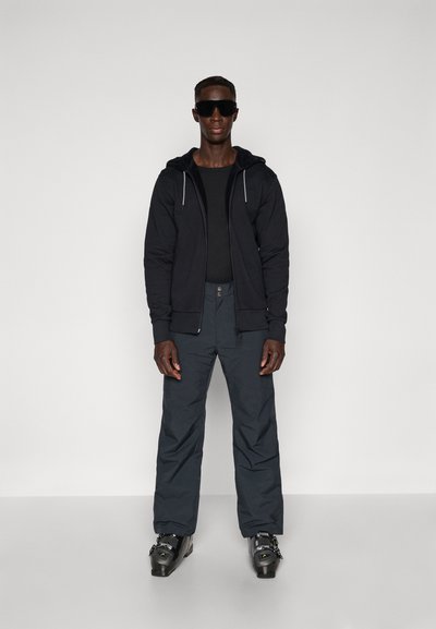 Haut à fermeture éclair noir sur une chemise noire, associé à un pantalon bleu marine. Les accessoires comprennent de grandes lunettes de soleil noires et des bottes noires avec des accents métalliques.
