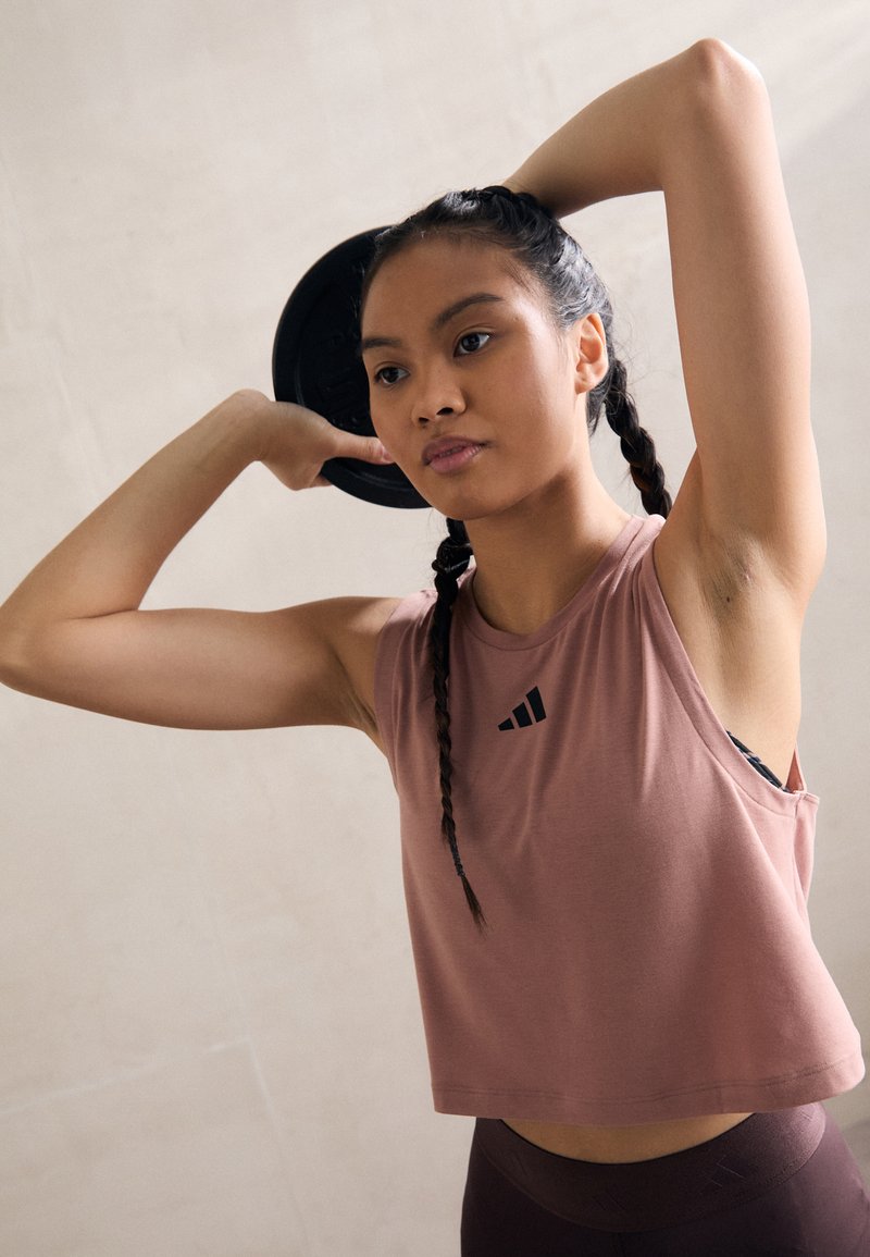 Crop top i støvet lyserød stof med sort logo; båret med mørke leggings. Motivet holder en vægtplade over hovedet og viser en atletisk kropsholdning.