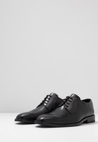 Scarpe da uomo in pelle nera con finitura liscia, punta rotonda, chiusura con lacci e tacco basso a blocco. Design minimalista senza ulteriori decorazioni.