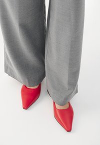 Red leather pointed-toe shoes with a smooth finish, paired with gray wide-leg trousers. The shoe has a low heel and a sleek design.