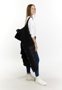 Sort lang frakke med dobbelt tekstur, hvor den nederste del er pufferet og den øverste glat. Model iført denim og hvide høj-top sneakers, stående foran en hvid baggrund.