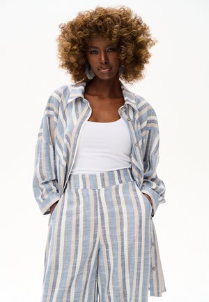 Kobieta z bujnymi kręconymi włosami nosi niebiesko-biało-beżową paskowaną kurtkę i spodnie z białym topem, ręce w kieszeniach, na białym tle.