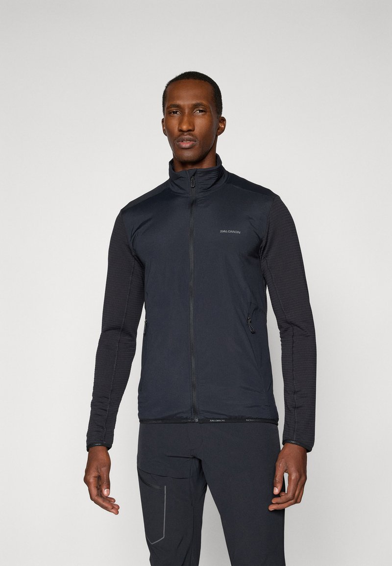 Un homme se tient de face, portant une veste zippée noire avec des poches latérales et un pantalon noir assorti, sur un fond gris uni.