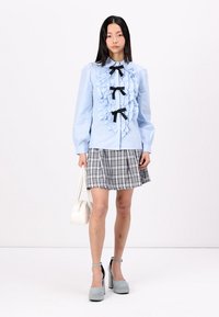 Light blue blouse with ruffled front, black bows, and long sleeves paired with a plaid mini skirt, accessorized with a white handbag and gray platform shoes.