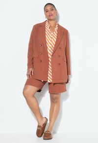 Hnědý dvouřadý blejzr přes oranžovo-bíle pruhované tričko, doplněný o hnědé šortky se stejným odstínem a hnědé nazouvací boty.