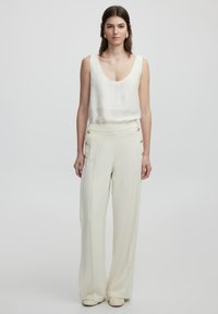 White sleeveless top with a rounded neckline and flowy fabric, paired with high-waisted cream trousers featuring gold button accents.