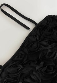 Černý textilní oděv s texturovanými květinovými aplikacemi, tenkými nastavitelnými popruhy a hladkým pozadím. Zahrnuje stříbrné kovové detaily.
