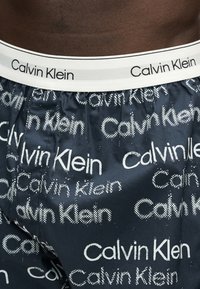 Κοντινή λήψη σκούρων μπλε boxer Calvin Klein με επαναλαμβανόμενα λευκά λογότυπα και λευκή μέση με την επωνυμία Calvin Klein.