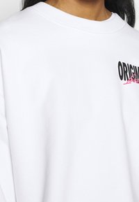 White sweatshirt made of cotton, featuring a black graphic text "ORIGINAL" and a pink accent, with a wide crew neckline and relaxed fit.