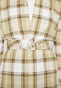 Sukienka koszulowa zapinana na guziki w brązowo-białą-zieloną kratkę, z szerokim paskiem z kwadratową klamrą i teksturą materiału.