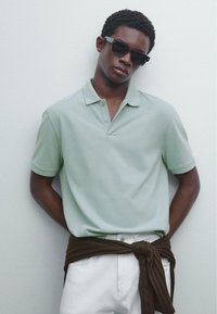 Ljusgrön polo med krage och tre knappar, tillsammans med vita byxor och ett brunt tyg knutet runt midjan.