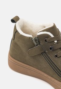 Botte en suede vert olive avec doublure en fausse fourrure blanche, fermeture éclair sur le côté et semelle en caoutchouc texturé. Comprend des lacets assortis et un col rembourré.
