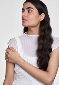 Ezüst karkötők egy csuklón, áttetsző, fehér, redőzött felsővel viselve, amelynek fodros nyaka van. Hosszú, sötét haj leomlik a vállra.