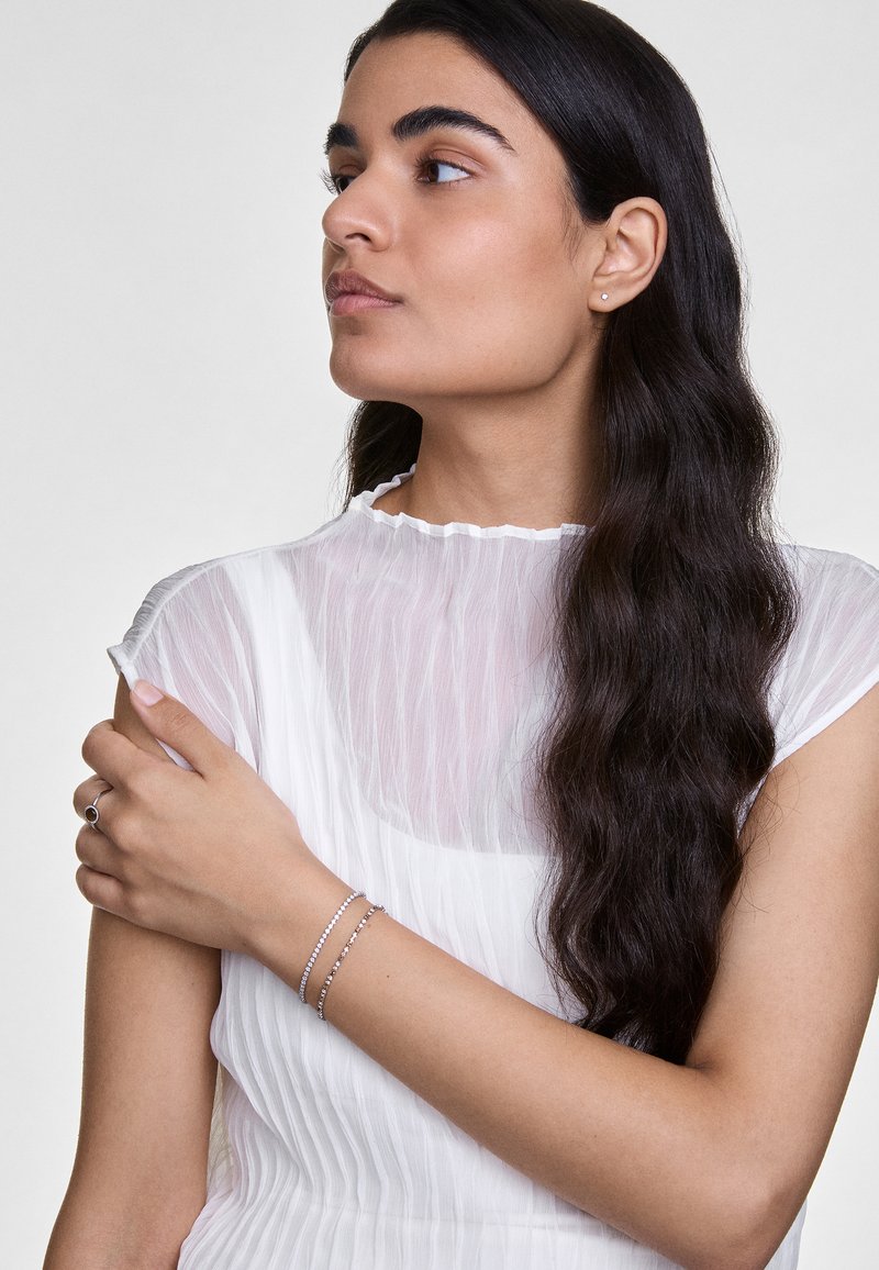 Ezüst karkötők egy csuklón, áttetsző, fehér, redőzött felsővel viselve, amelynek fodros nyaka van. Hosszú, sötét haj leomlik a vállra.