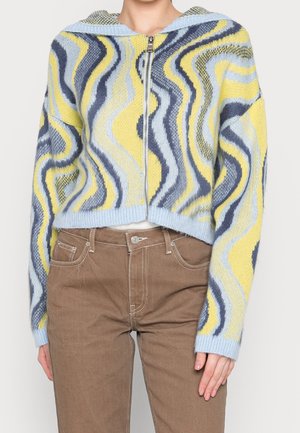 Personne portant un pull zippé en tricot avec des motifs ondulés jaunes, bleus et gris, associé à un pantalon marron taille haute.