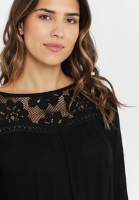 Svart blus med spetsöverdrag med blommönster längs halsringningen, långa ärmar och en lös, flödande passform.