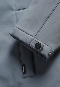 Svetlo siva jakna srajcni rokav in gumbom ter dodatnimi detajli. Gladka tekstura z vidno etiketo z napisom "Martinique." Posnetek od blizu.