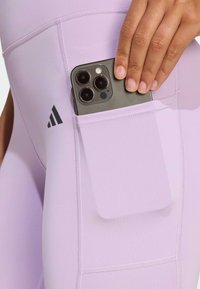 Leggings violets avec une poche contenant un smartphone, présentant un design sans couture et une texture subtile, soulignés par des accents de logo noir.