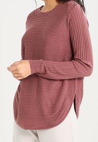 Frau, die einen pulvrigen rosa gerippten Strickpullover mit langen Ärmeln und Seitenschlitzen trägt, kombiniert mit hellen Hosen.