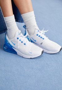 Feet wearing white and light blue Nike Air 270 sneakers with white socks, resting on a light blue carpet.