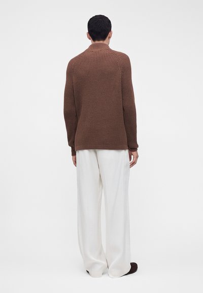 Personne de dos portant un pull à côtes marron et un pantalon large blanc sur un fond blanc uni.