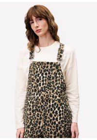 Vrouw met golvend bruin haar, gekleed in een crème lange mouw shirt en een luipaardprint tuinbroek, staand tegen een effen witte achtergrond.