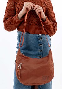 Bruine gestructureerde schoudertas met rits, een klein buitenzakje en verstelbare banden, vastgehouden door iemand die een gebreide trui draagt.