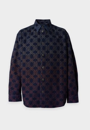 Homme portant une chemise boutonnée sombre à motifs, jean bleu, baskets blanches, debout devant un fond clair uni.