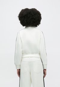 Λευκό κοντό φούτερ με μαύρες ρίγες στο πλάι και ριμπ μανσέτες. Συνδυάζεται με ανάλογο ελαφρύ παντελόνι με πίσω τσέπες.