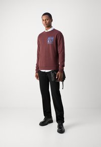 Felpa maroon con grafica blu, pantaloni neri e scarpe nere. Tiene una piccola borsa nera con cinghie. Sfondo semplice.