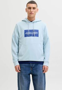 Jeune homme portant un sweat à capuche Jack & Jones bleu clair et un jean bleu, debout devant un fond blanc uni.