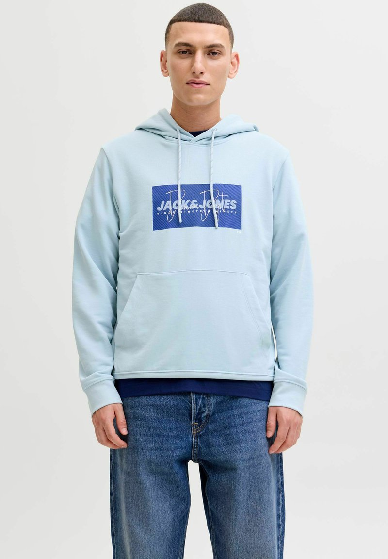 Jeune homme portant un sweat à capuche Jack & Jones bleu clair et un jean bleu, debout devant un fond blanc uni.