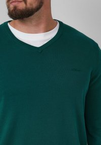 V-Ausschnitt Pullover in Dunkelgrün, aus Strickstoff gefertigt. Verfügt über ein dezentes Logo auf der linken Brust und ein kontrastierendes weißes Innenhemd.