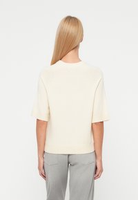 Femme aux longs cheveux blonds portant un pull crème à manches courtes et un jean gris clair, vue de dos sur un fond blanc.