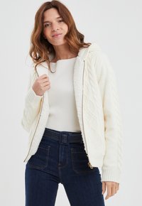 Giacca con cappuccio color crema con design a treccia testurizzato, fodera in pile e polsini a coste, abbinata a un top bianco a coste e jeans di denim scuro.